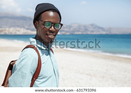 Attractive fashionable black man tourist dressed in trendy clothing and accessories posing against blue water and white sand background with copy space for your information during vacations at seaside