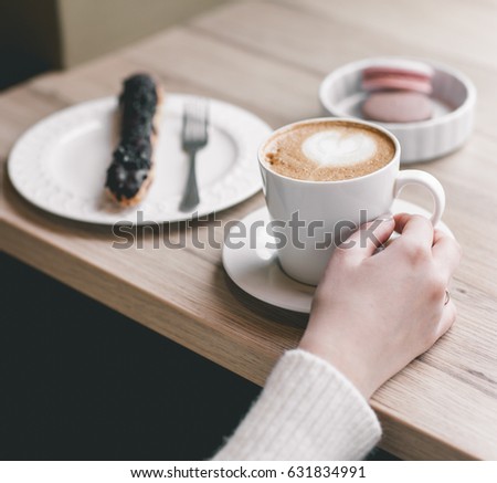 Womans hands wrapped around a cup on wood table with chocolate eclair