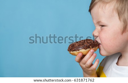 Little happy cute boy is eating donut on blue background wall. child is having fun with donut. Tasty food for kids. Funny time at home with sweet food. Bright kid.