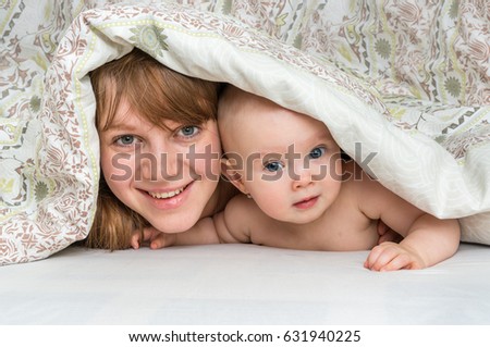 Mother and her baby playing and smiling under a blanket in bed  - happy family concept 