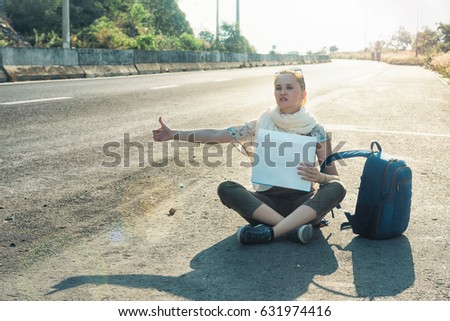 Young woman hitchhiker sitting on the road. Tired girl with blue backpack holding a white blank cardboard sign. Hitchhiking at sunset or sunrise