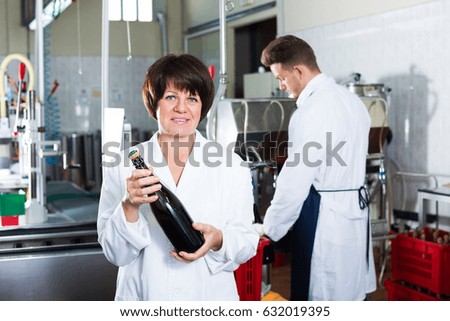Positive senior woman worker displaying wine bottle at sparkling wine factory