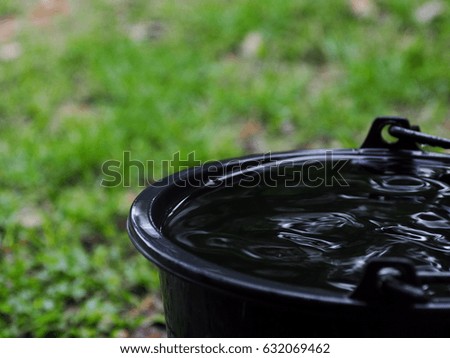 dark gray plastic bucket on the floor outdoor surrounding with green area environment with reflection on water waves surface in the bucket