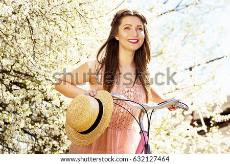 Flower portrait of tender, smiling young and beautiful long-haired girl in pink dress riding a bike and keeping hat on flower background.
