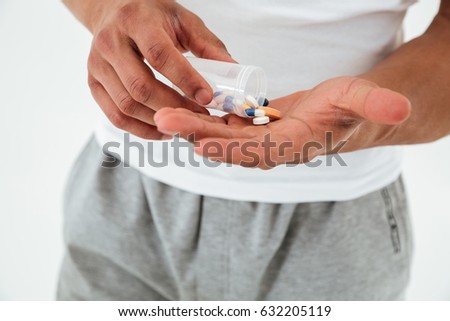 Cropped picture of young sportsman standing over white background holding vitamins and sport pills.