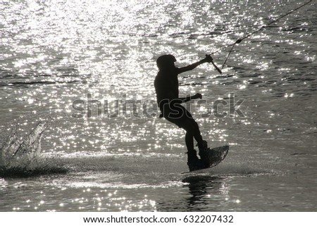 Wake Boarder Being Pulled by Cable Slightly Elevated in Mid-Air Left to Right in Silhouette Late Afternoon against Sunlit Water Sparkles in Ski Rixen Lake, Quiet Waters Park, Deerfield Beach, Florida