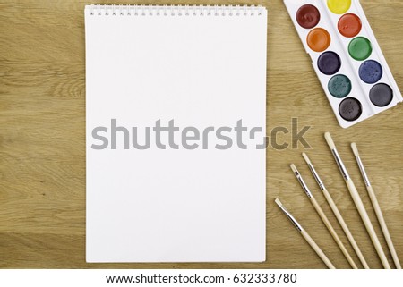 Set of watercolor paints, brushes and blank white paper sheet on wooden background. Top view.