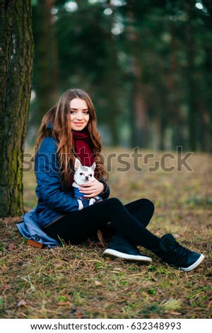 Young and lovely girl with long hair in blue jacket sitting on ground beyond trees in forest with her lovely chihuahua puppy in dog clothes. Pet care. Innocent domestic animal. Nature. Pine. Friends.