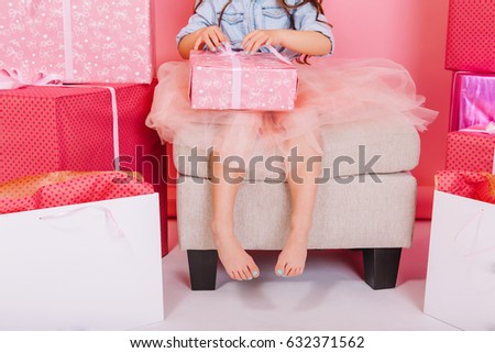 Lovely cute  image of birthday kid opening present on knees suround big giftboxes, balloons on white floor. Amazing little princess in tulle skirt celebrating birthday party