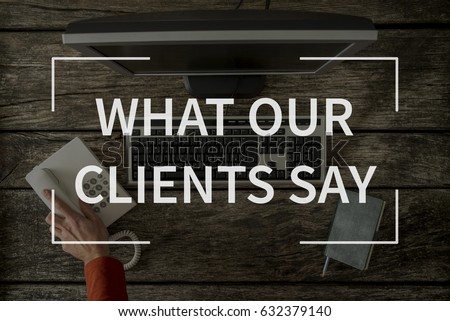 What our clients say text over top view image of hand reaching for phone seated in front of computer keyboard, monitor and notepad. Royalty-Free Stock Photo #632379140