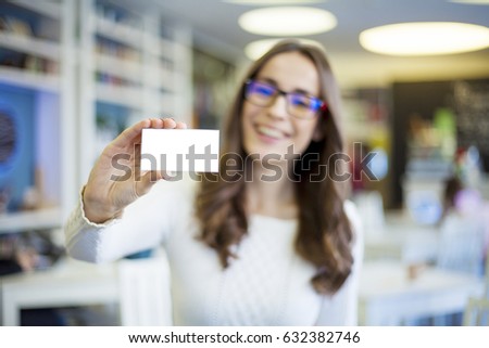Portrait of attractive smiling brunette business woman in glasses with visit card in hand on the modern startup office background.