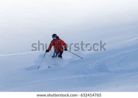 Skier in orange jacket and black helmet making turns in snow powder with mountains in the background