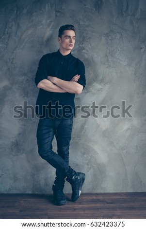 Young attractive model in black shirt, jeans and stylish boots, nice hairstyle, is standing on grey background, he crossed his arms and looks at his left Royalty-Free Stock Photo #632423375