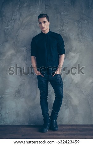 Young attractive model in black shirt, jeans and stylish boots standing on grey background, he put his hands in the pockets Royalty-Free Stock Photo #632423459