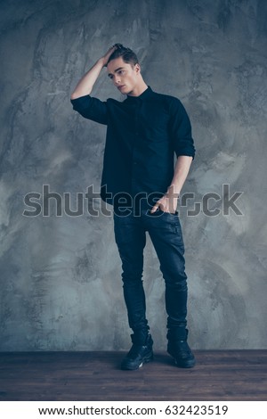Young handsome fashion man model in black shirt, jeans and stylish sneakers posing on grey background, touching his hair Royalty-Free Stock Photo #632423519