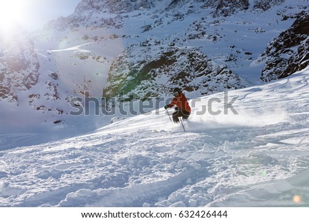 Sun flared skier in orange jacket making turns in fresh powder snow with mountains on the background.