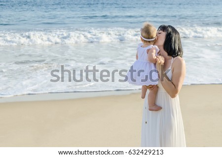 Mother is kissing her baby girl on the sandy beach near ocean in sunny day/Mothers day concept