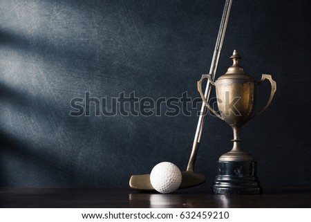 still life photography :  golf club (putter) and ball with old trophy on wood table against art dark background with window light at left