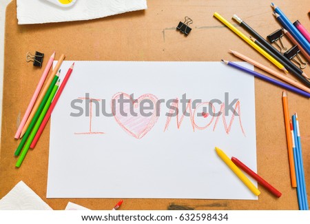 Sketch paper with a sentence "I love mom" surrounding by pencils, painting brush, tray of white, yellow, blue, red, green, yellow colour and black clips over cork board in art class, top view