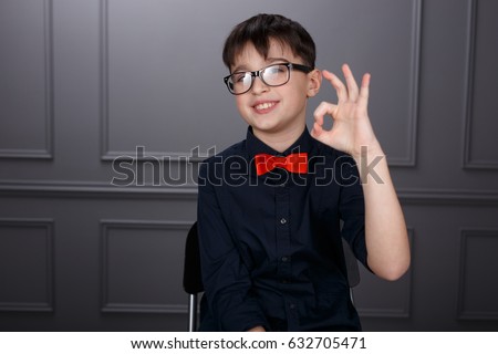 Amusing smiling brunette young boy in black shirt and red bow tie, in eyeglasses, showing ok hand sign, isolated on grey wall background. Horizontal view. 