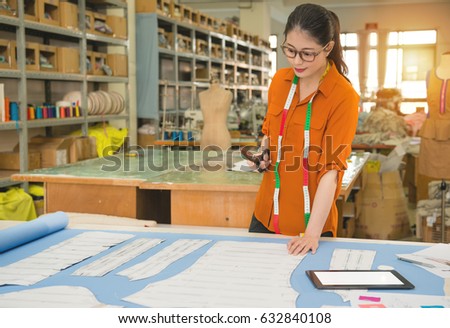 smile elegant clothing tailor standing at desk holding scissors thinking about pattern how to design new clothes prepare for new season with blank screen mobile computer in factory office.