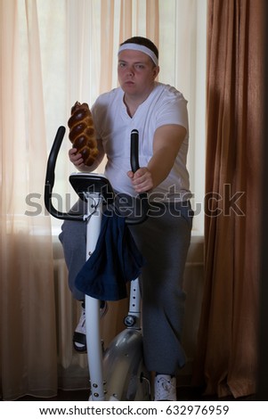 Fat guy exercising on stationary training bicycle and eating a bun at home