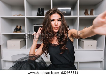 Happy gorgeous young woman with curly brown hair, dancing and taking selfie in her dressing room. She showing kiss to camera. She enjoying good day. Wearing black dress.