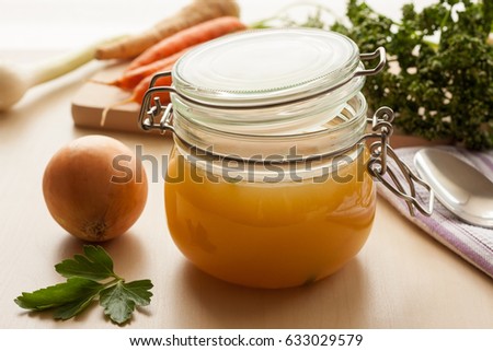 Bone broth made from chicken in a glass jar, with carrots, onions and parsley in the background
