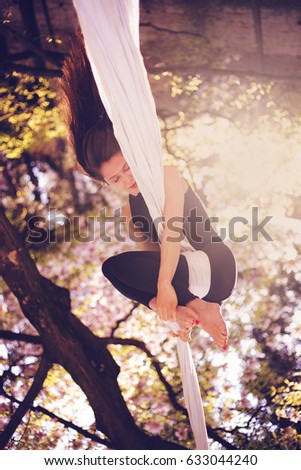 Beautiful girl in dress performing aerial silk dance on the white ribbons in sunny spring nature under the trees with pink flowers in the park on a sun
