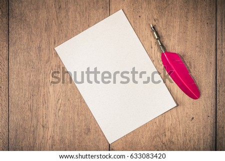 Vintage red quill ink pen and textured sheet of paper blank on oak wooden desk. Retro style filtered photo
