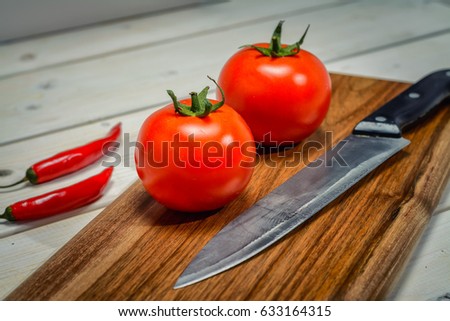 Two red tomatoes and kitchen knife on a cutting board next to two spicy peppers