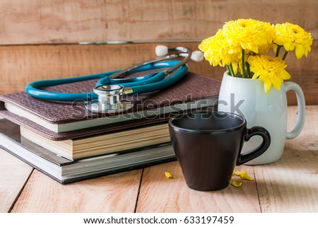 cup of hot coffee with stethoscope on books and yellow flower in white pot on wooden table.
