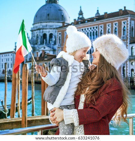 Another world vacation. smiling trendy mother and child travellers on embankment in Venice, Italy in the winter with Italian flag
