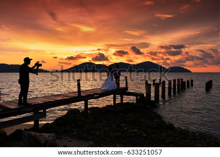 Silhouette photographer taking photo of wedding couple on the wooden jetty with beautiful sunset background.