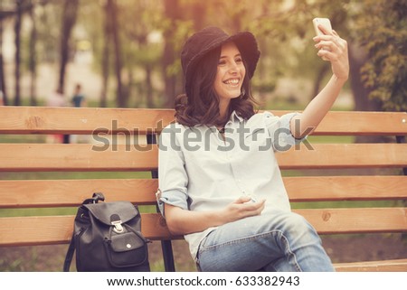 a beautiful woman is taking a selfie picture while in the park, using her mobile phone, seating at a bench