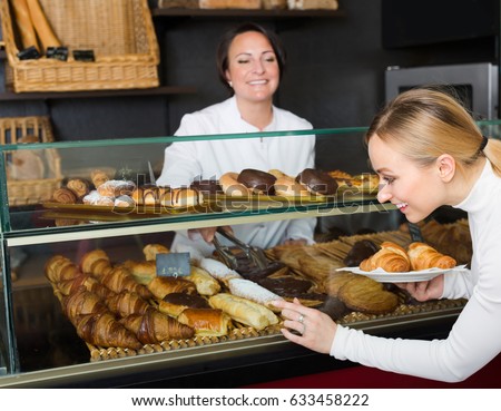 Smiling mature woman dressed as cook showing deserts in the pastry shop. Selective focus on customer
