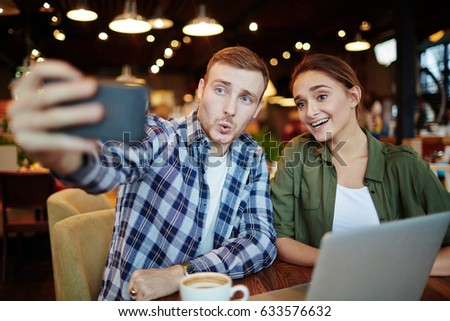 Having good time in lovely coffeehouse: handsome young man showing grimace while taking selfie with his smiling pretty friend, waist-up portrait