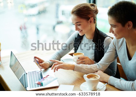 Profile view of pretty short-haired woman sitting at cafe table with her smiling friend and showing her photos from vacation, waist-up portrait