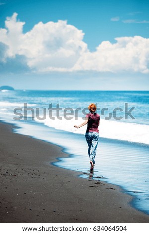 A tall happy caucasian girl running on a  beach along the surf line. Tropical beach with black volcano sand and blue sky with clouds. Happiness and freedom concept. Vertical photo. Bali, Indonesia