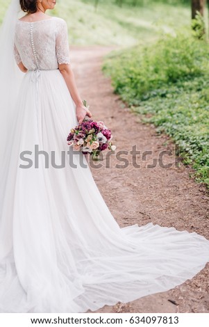 Bride is standing in forest with a bouquet in her hand showing the back of her white wedding dress and it hemline. Wedding. Details