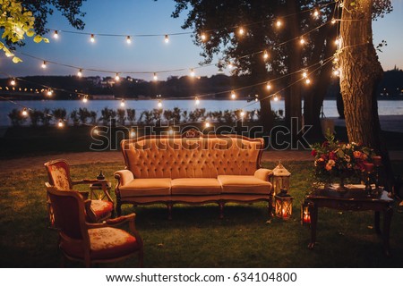 Wedding. In the forest on the river bank there is sofa and armchair, next to it there is wooden table with composition of flowers and greens. candles on the grass. hang garlands of light bulbs.