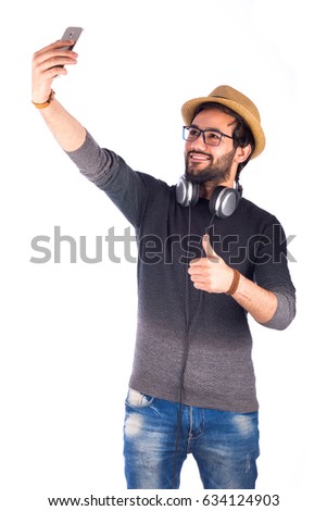 Happy friendly young man with glasses taking selfie and giving thumb up, guy wearing gray t-shirt and jeans with headphone and beige hat, isolated on white background