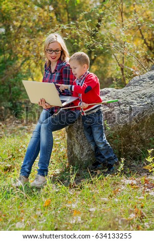 young beautiful mom with glasses is holding a laptop on her hands and her son is  showing something in the laptop, with a self-stick in his hands. 