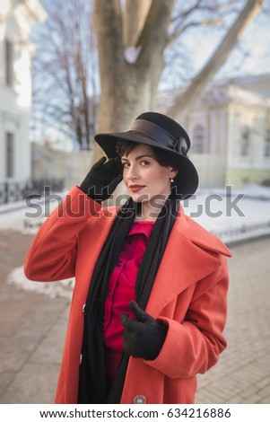 The lovely girl in a red coat, a hat and a crimson dress. She attractively looks at us. The girl has a gentle smile and red lips.