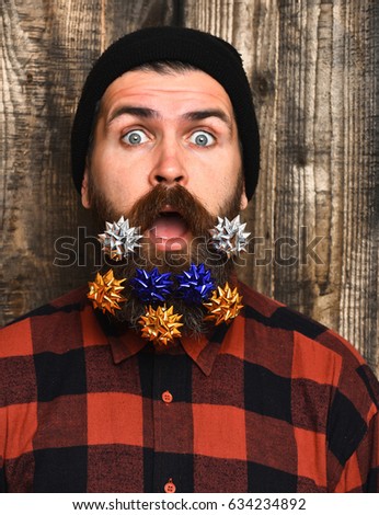 Bearded man, long beard. Brutal caucasian surprised hipster with colorful gift decoration stars in moustache, wearing red black checkered shirt on brown wooden vintage studio background