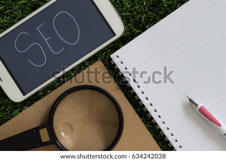 Wood Letters "SEO" abbreviation (Search Engine Optimization) on Smart Mobile Phone Screen with Magnifying Glass and Pen on Blank Notebook. Idea Concept for Organization, Business System.