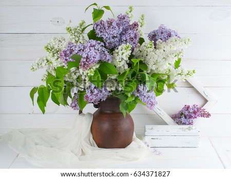 Lilac bouquet in clay jug with tulle fabric, vintage casket and retro photo frame on background of shabby wooden planks in rustic style 