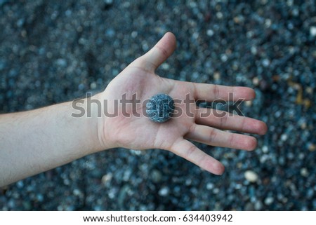 Looking down from above onto an outstretched arm and hand holding a small, round pebble with a course, stony background 