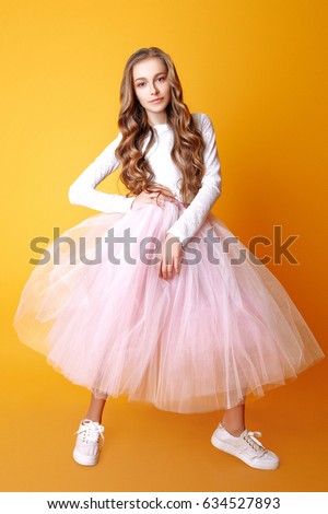 beautiful teenage girl with long curly hair on a yellow background. Studio photo. Dressed in a white sweater and pink tulle full skirt