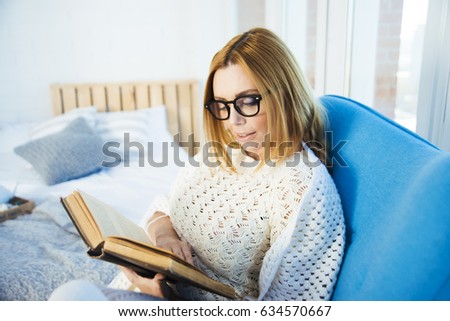 Beautiful blonde woman in white sweater sit on couch wearing glass reading big book. interior living room. day light